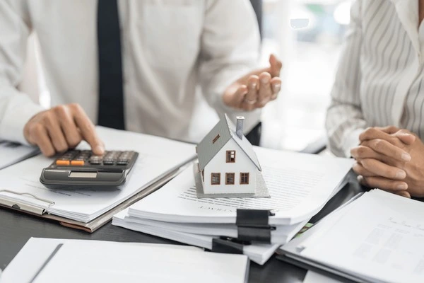 Man using a calculator in front of a pile of paperwork with a small model house